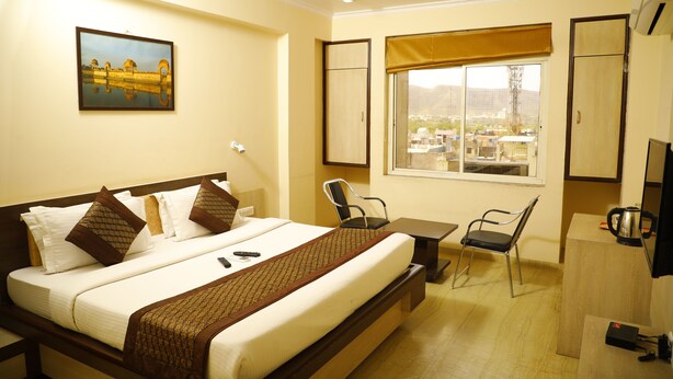 A hotel room with a bed, a desk, a chair, a television, and a window with a view of buildings. - The Prime Hotel Jaipur (Jaipur)