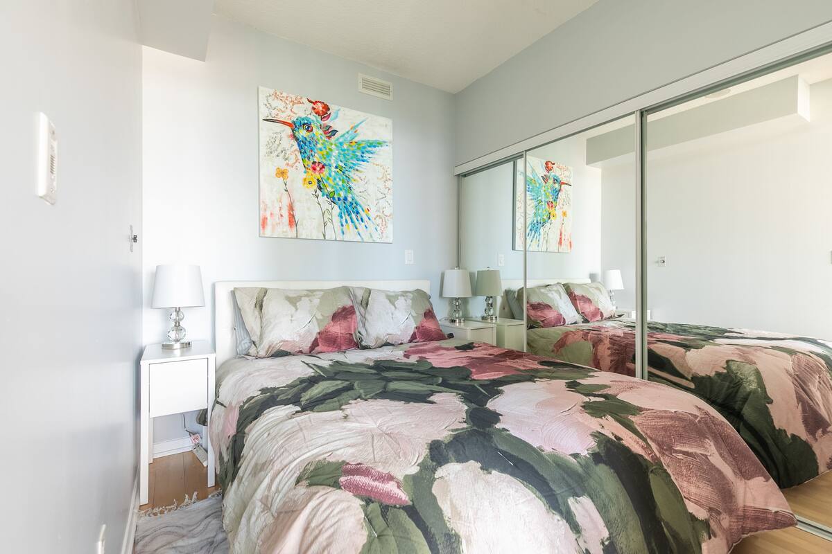Un dormitorio con una cama, una mesita de noche, una lámpara, un espejo y una pared con un cuadro floral.