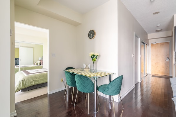 Un coin repas avec une table en verre et quatre chaises vertes, une horloge au mur et une vue sur une chambre à coucher avec un lit et une table de chevet.