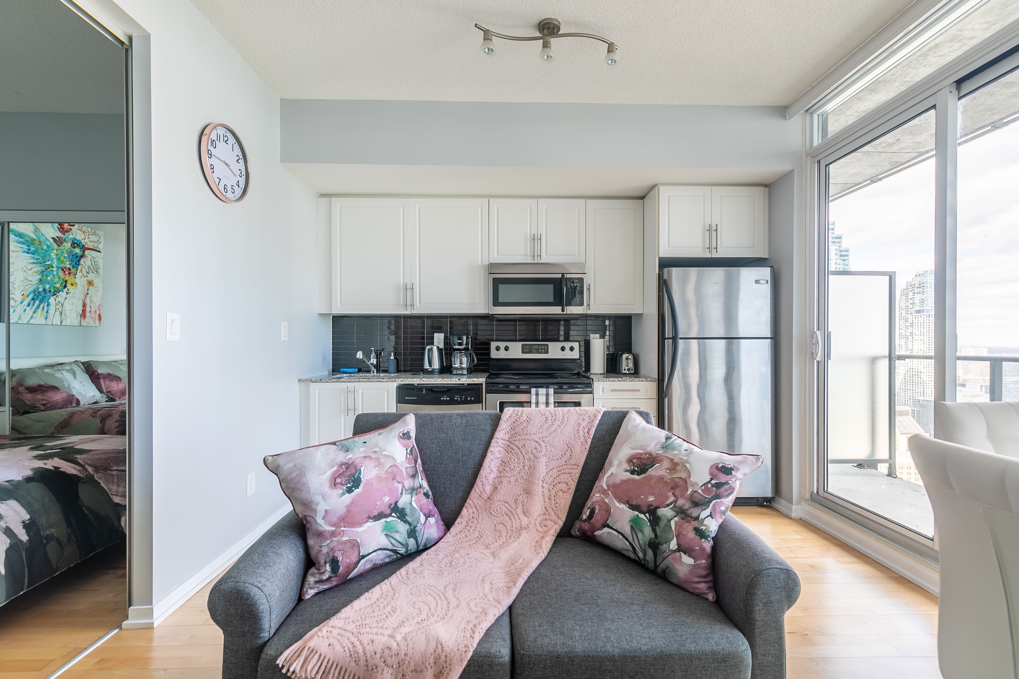 A modern apartment with a grey sofa, floral cushions, a pink throw, a kitchen with white cabinets, a stainless steel refrigerator, and a large window offering a city view.