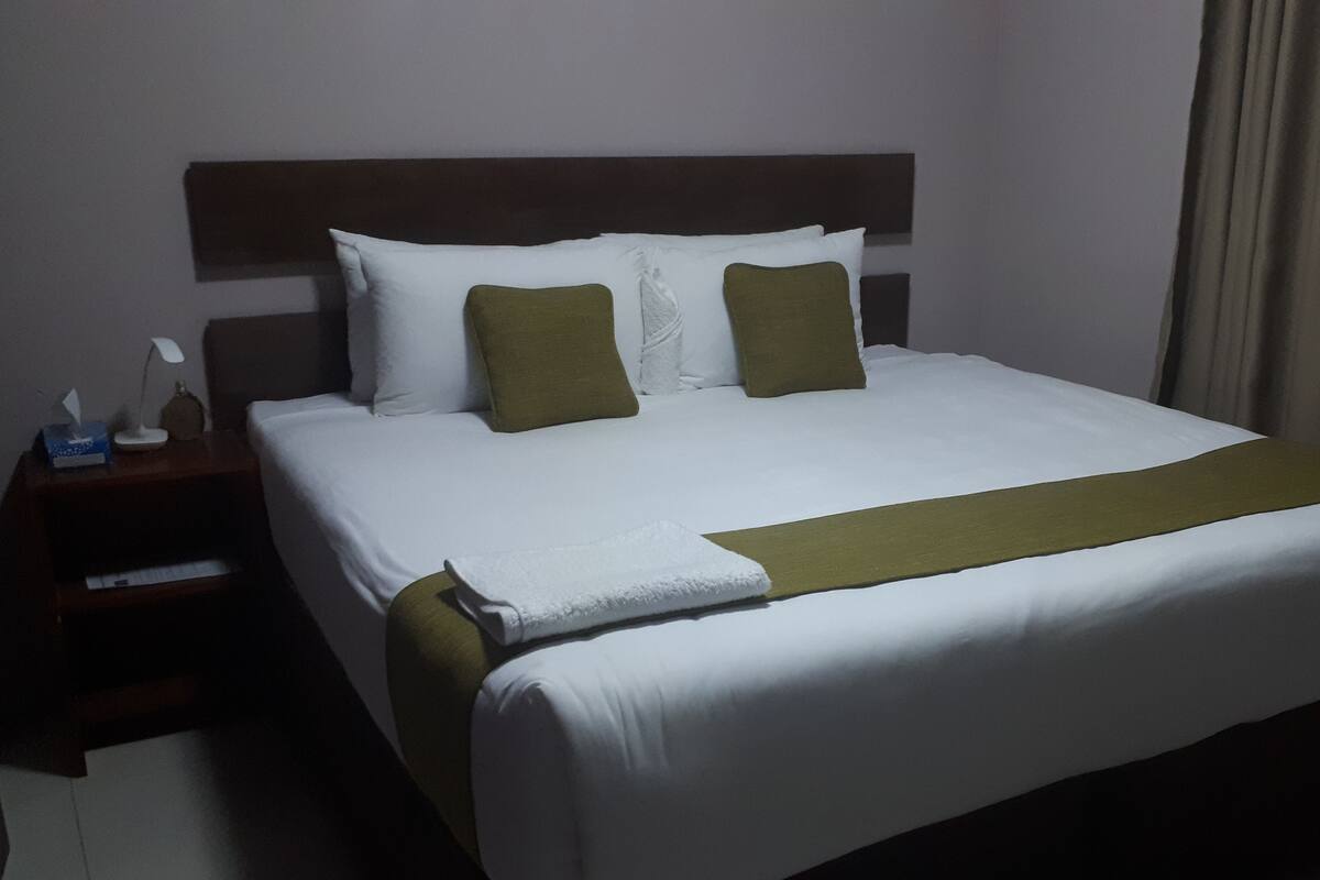 A neatly made bed with white bedding and two green pillows, a wooden headboard, a bedside table with a lamp and tissues, and a beige curtain.