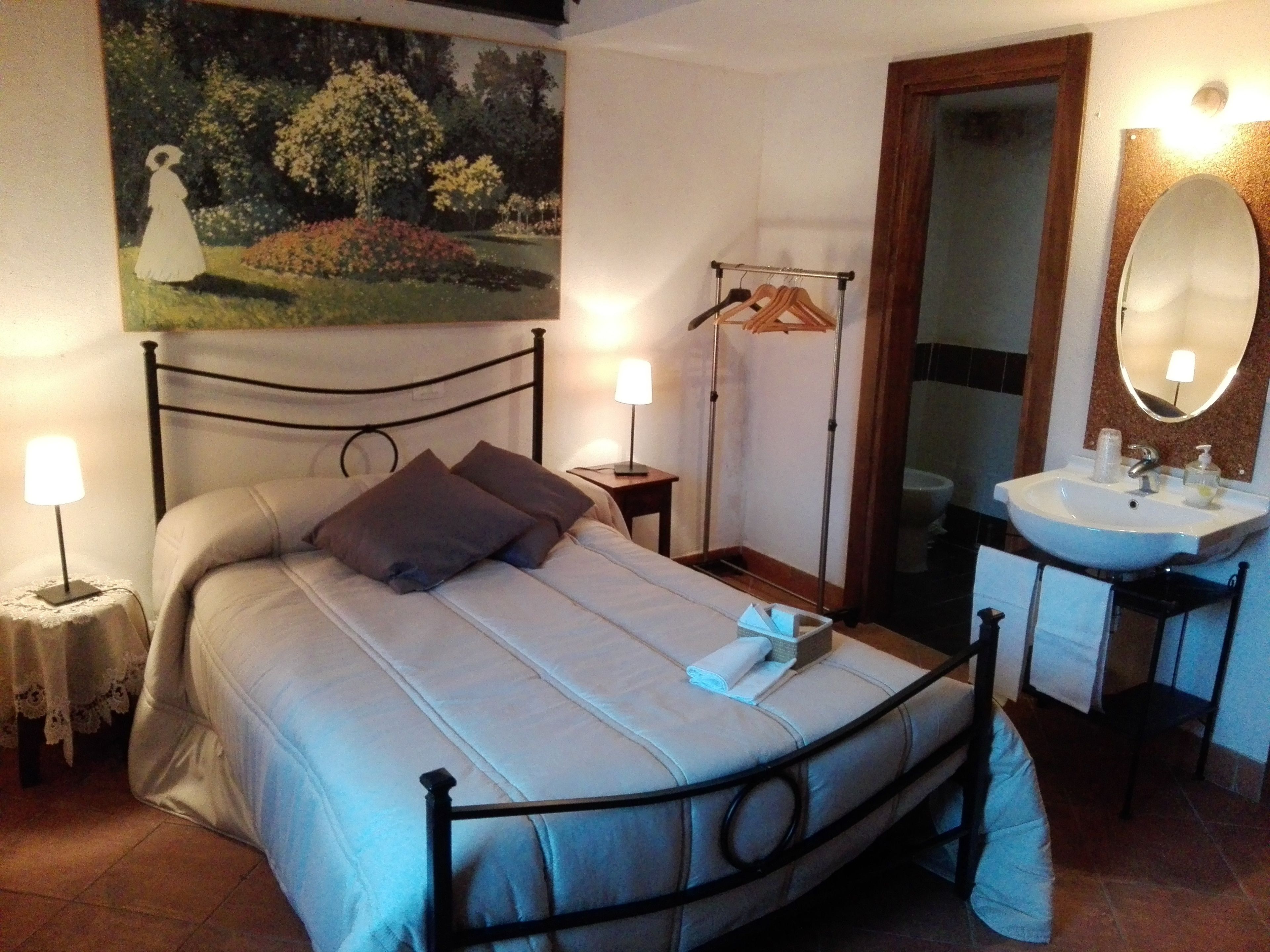 A bedroom with a bed, bedside lamps, a painting, a wooden hanger, a bathroom with a sink and mirror.