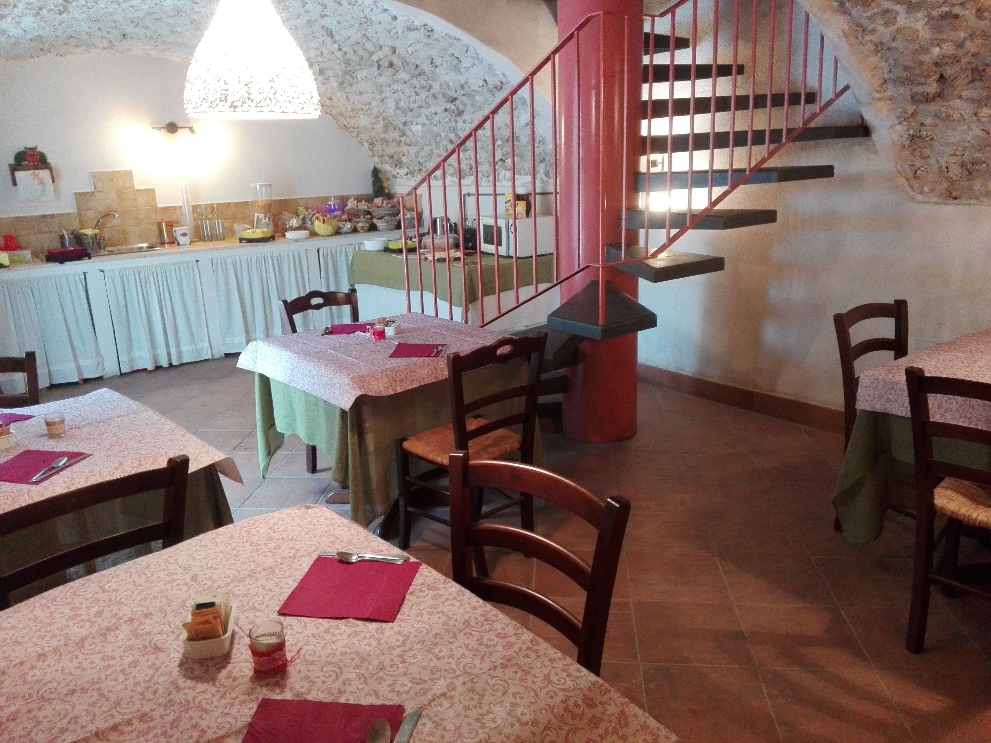 Zona pranzo con tavoli apparecchiati per due, una scala e una cucina sullo sfondo.