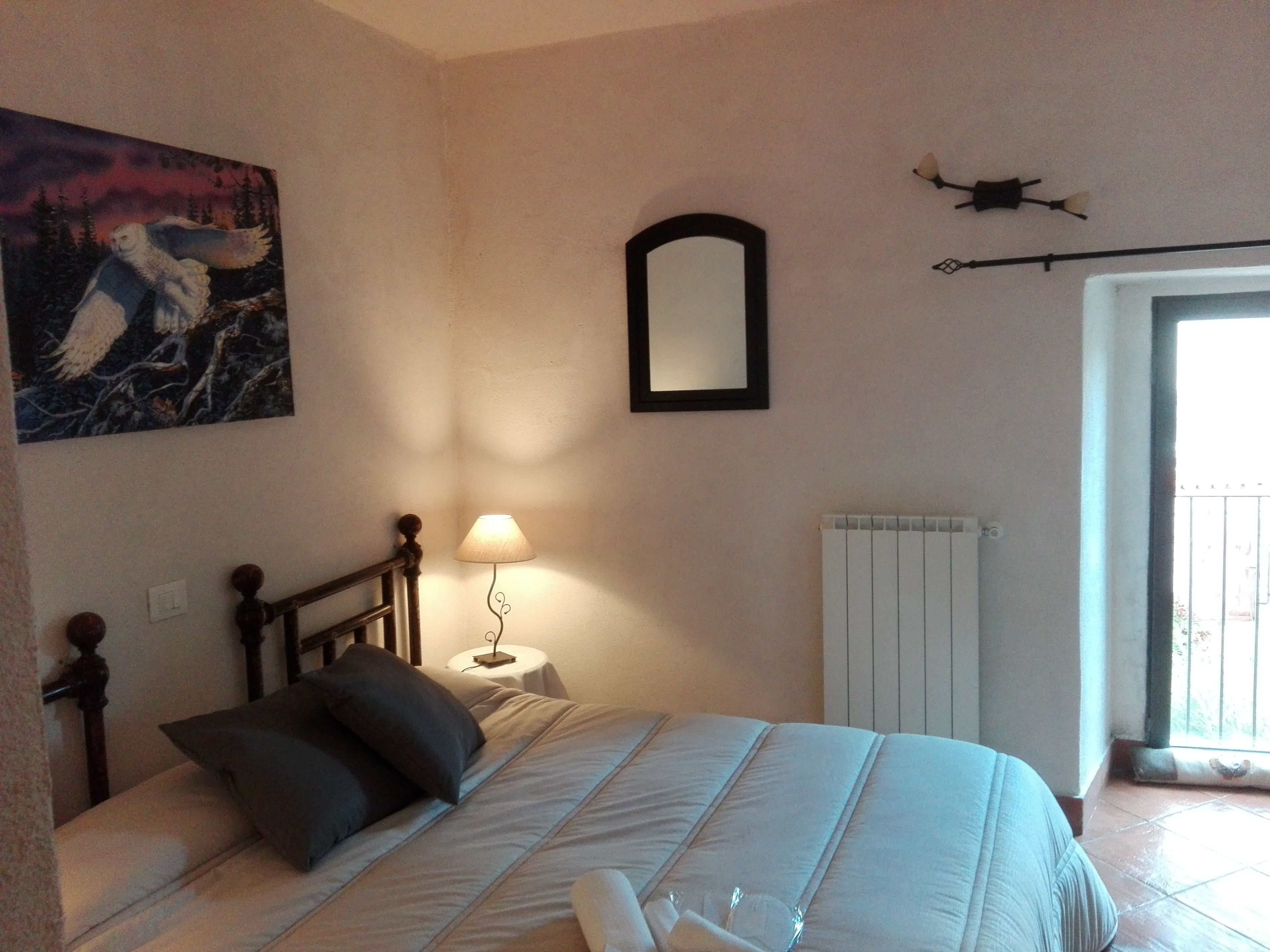 Un dormitorio con cama, mesita de noche, lámpara, cuadro, espejo y ventana.