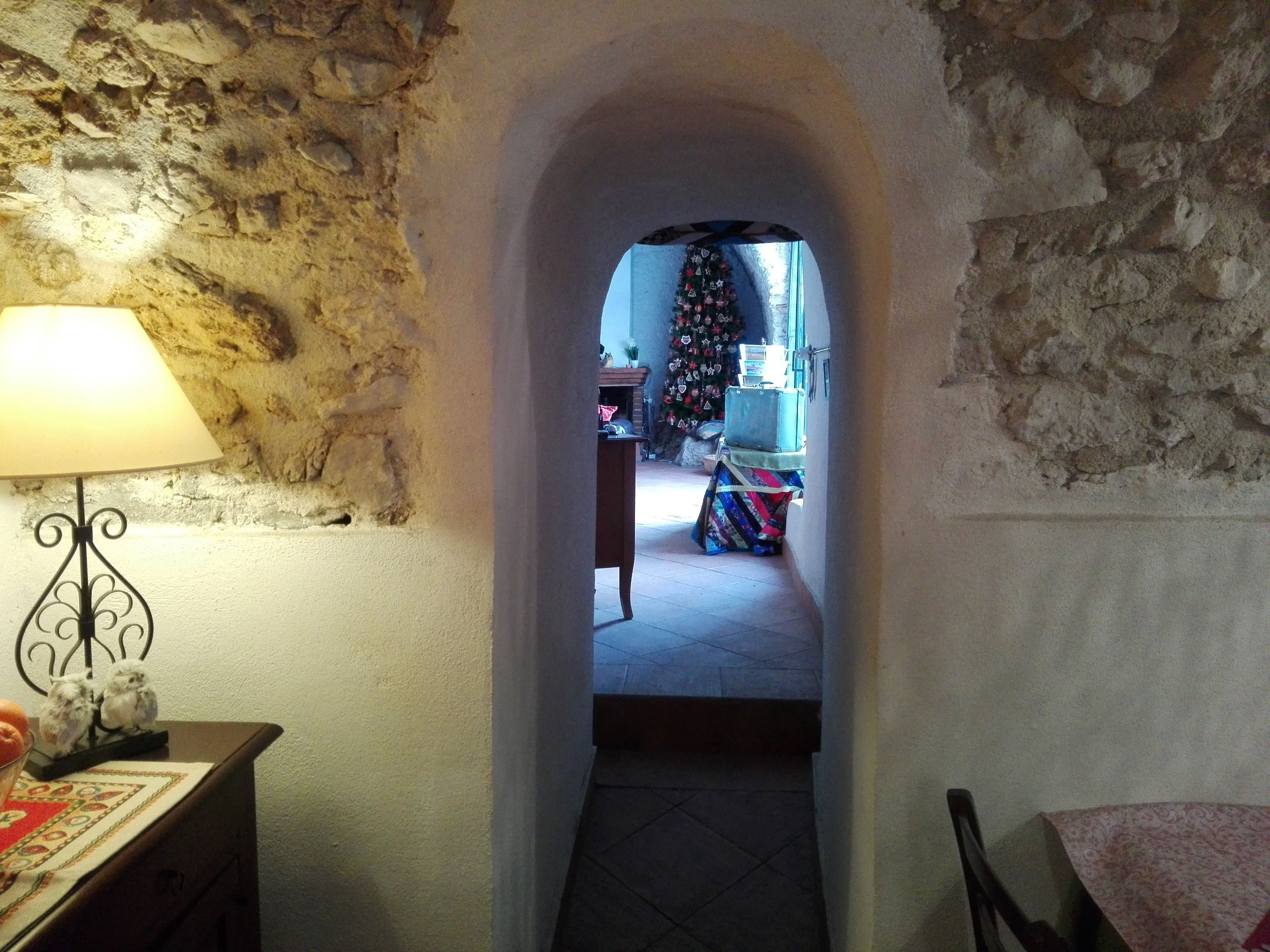 Un estrecho pasillo de piedra que conduce a una habitación con un árbol de Navidad, una lámpara y una mesa con mantel estampado.