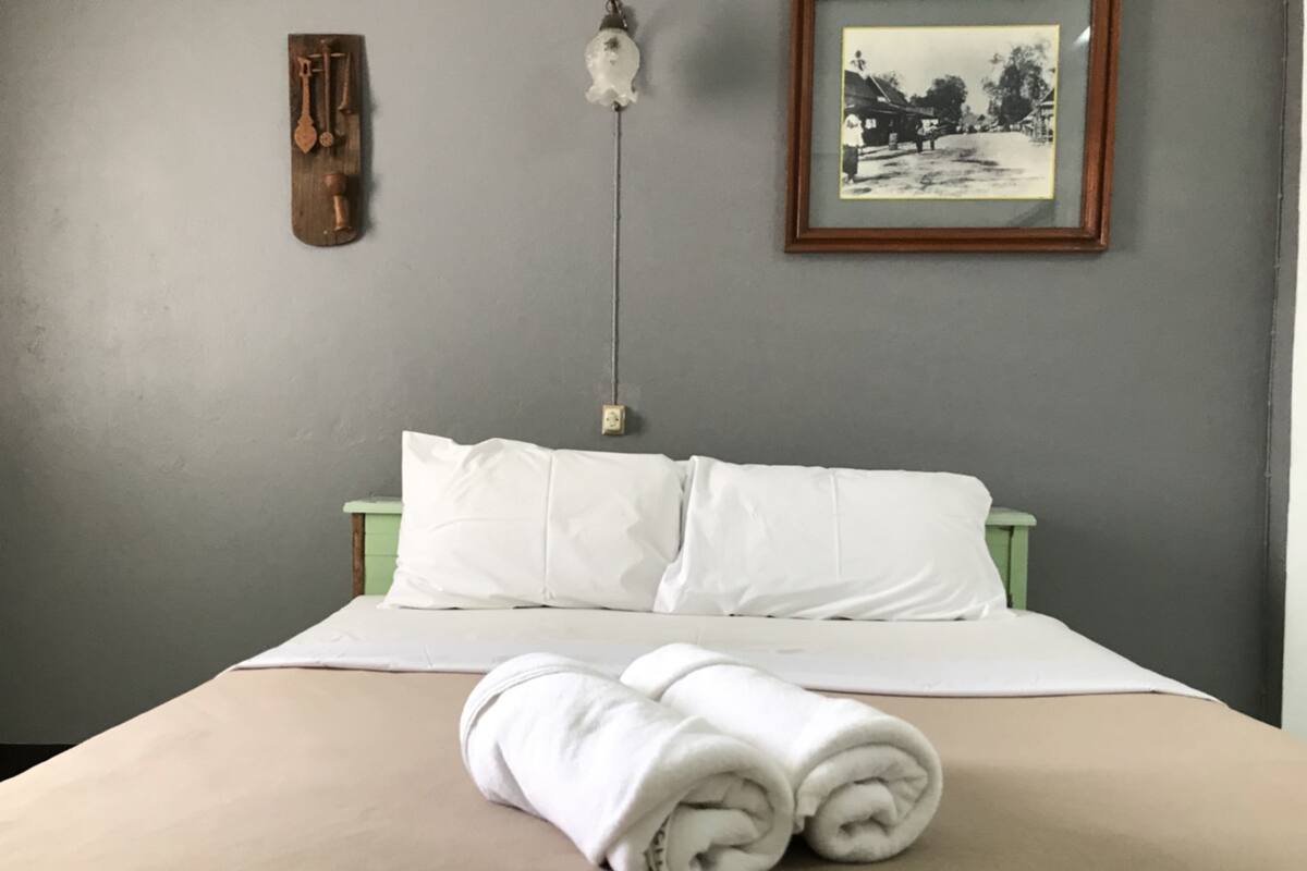Een net opgemaakt bed met witte lakens, een opgerolde handdoek, een zwart-wit foto in een lijst en een wandlamp.