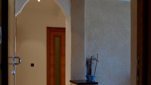 Un pasillo con una puerta de madera, un pequeño jarrón azul sobre un pedestal, y un suelo de baldosas.