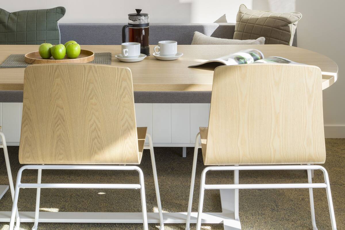 Uma sala de jantar com uma mesa de madeira, duas cadeiras, uma planta emoldurada, uma cafeteira e uma tigela de maçãs.