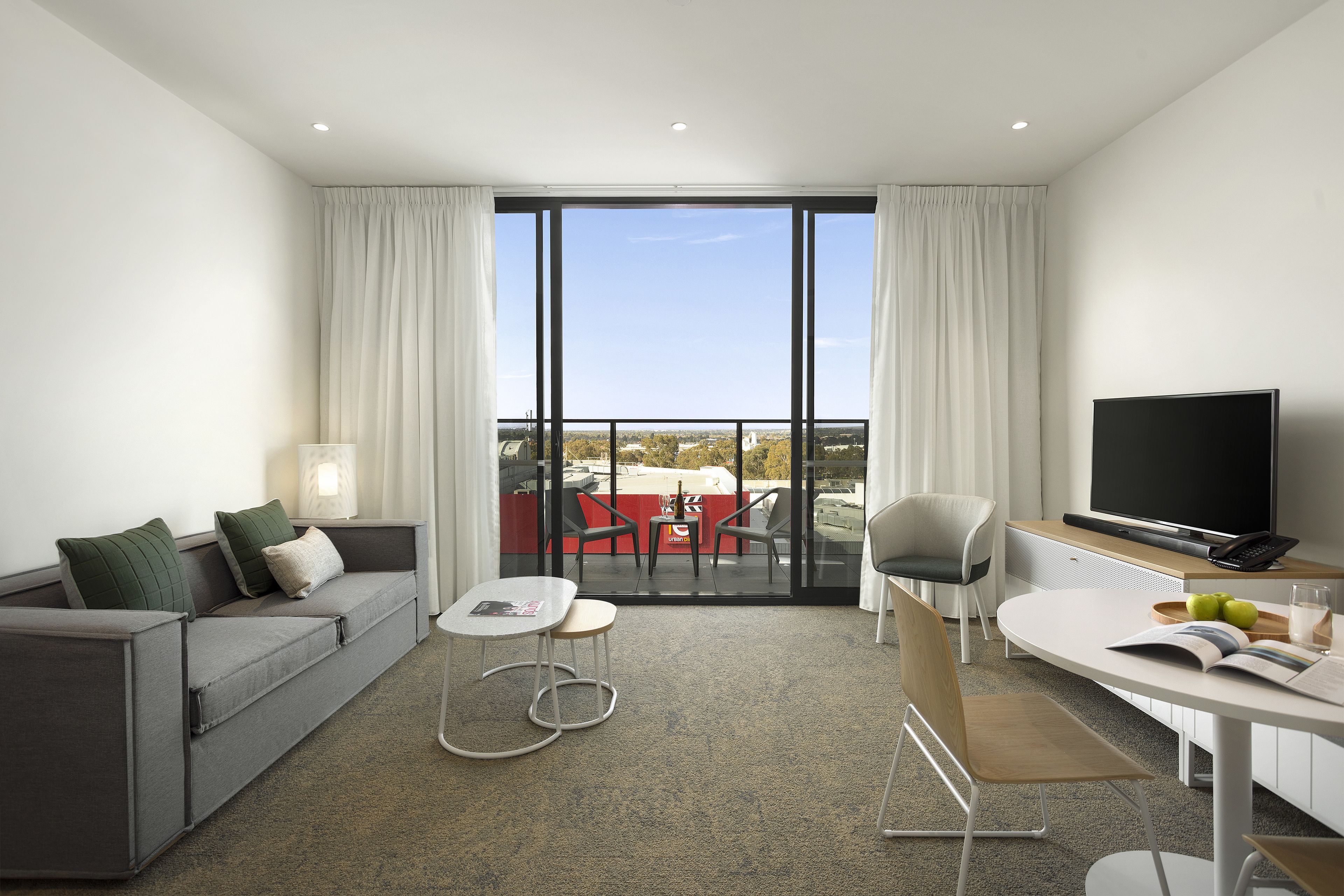 A modern hotel room with a grey sofa, a small table, a TV, and a balcony with a red table and chairs.