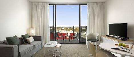 Habitación de hotel moderna con un sofá gris, una mesita, un televisor y un balcón con una mesa y sillas rojas.