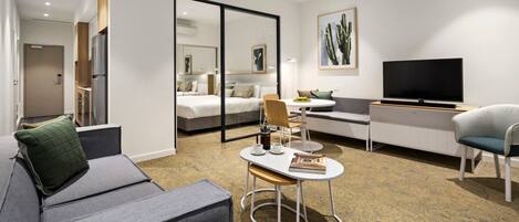 A modern hotel room with a sofa, a small table with a book and a cup, a TV, a bed, and a large mirror.