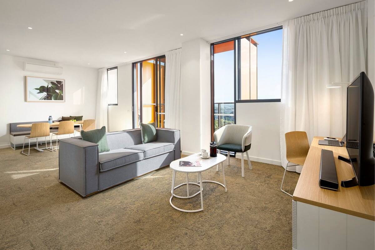 Quarto de hotel moderno com um sofá cinza, uma pequena mesa branca, uma escrivaninha de madeira e uma televisão.