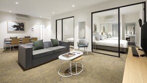 A modern hotel room with a grey sofa, a small white table, a bed with a grey headboard, a TV, and a large mirror. - Quest Epping (Epping)