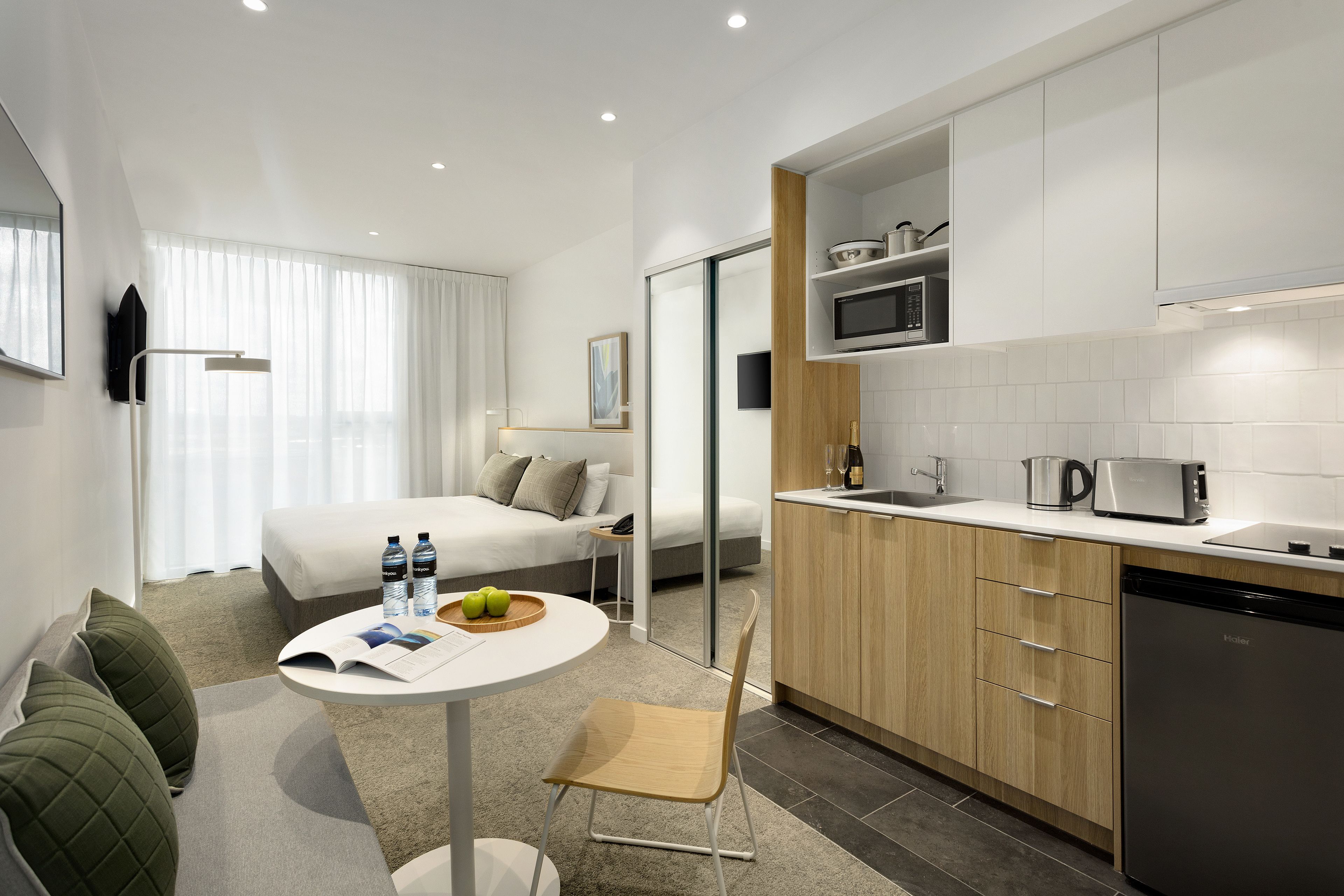 A modern hotel room with a bed, a small round table with a magazine and two water bottles, a chair, a kitchenette with a microwave, and a coffee maker.