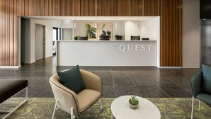 A modern reception desk with the word "QUEST" prominently displayed, surrounded by a wooden panel wall, a reception area, and seating arrangements. - Quest Epping (Epping)
