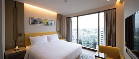A hotel room with a large bed, a bedside table with a lamp, a chair, a large window with curtains, and a city view.