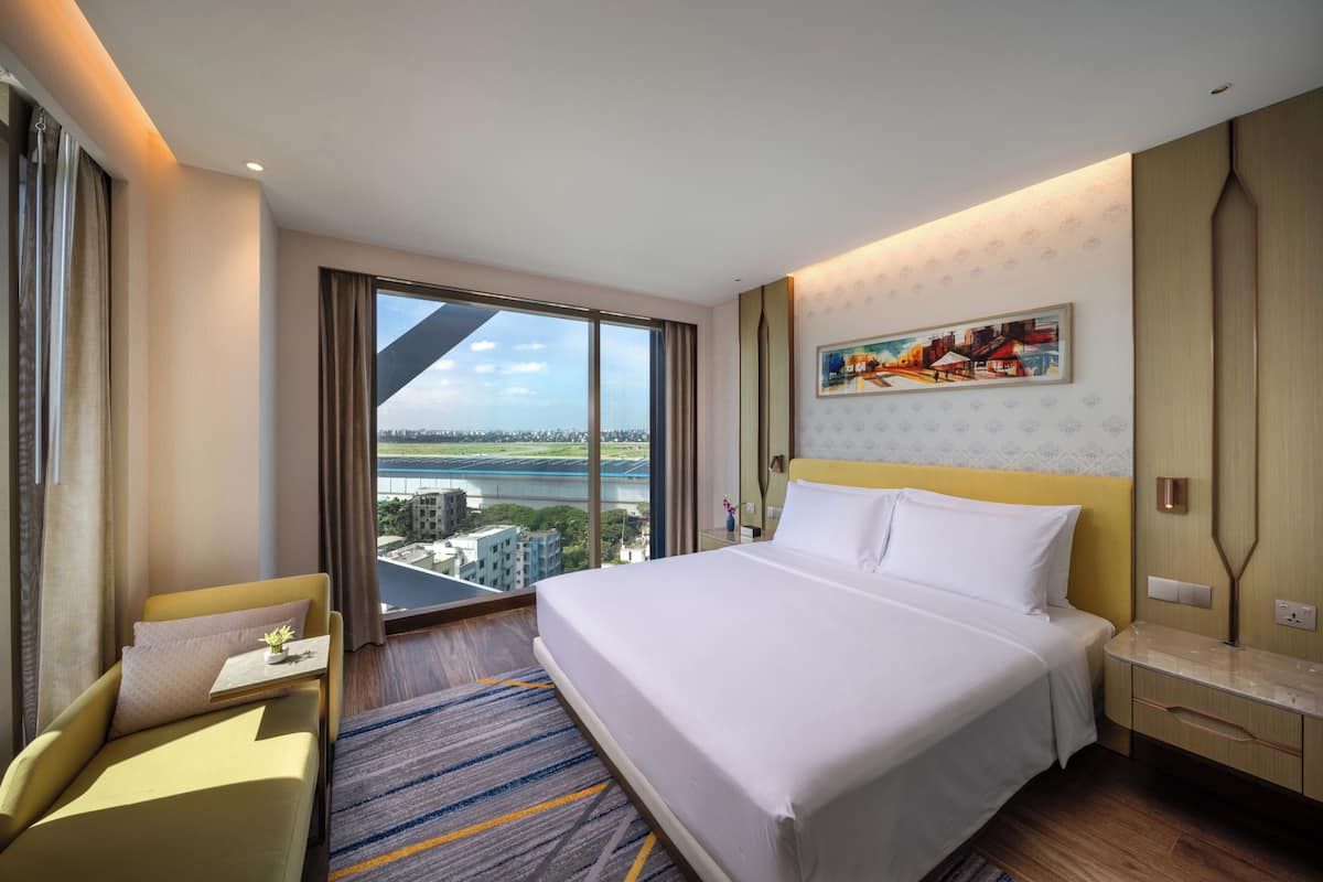 A hotel room with a large bed, a sofa, a nightstand, and a view of a cityscape through large windows.