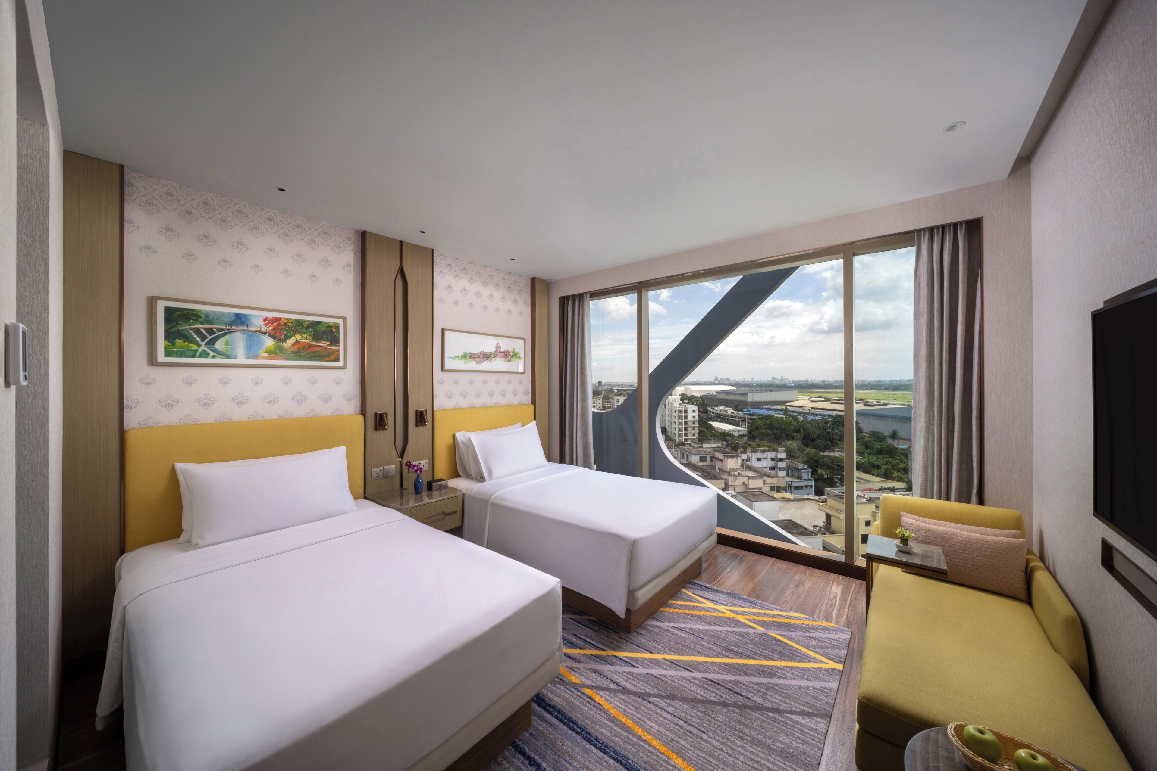 Habitación de hotel con dos camas, un sofá, un ventanal grande con vistas a la ciudad y obras de arte en las paredes.