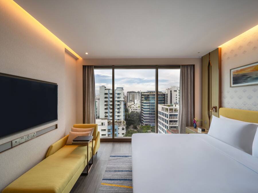 Quarto de hotel com cama grande, sofá, televisão e vista para a paisagem urbana.