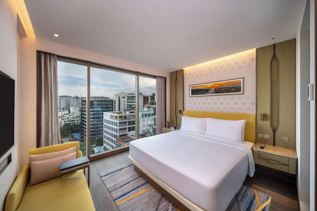 A hotel room with a large bed, a sofa, a desk, and a view of the city through large windows.