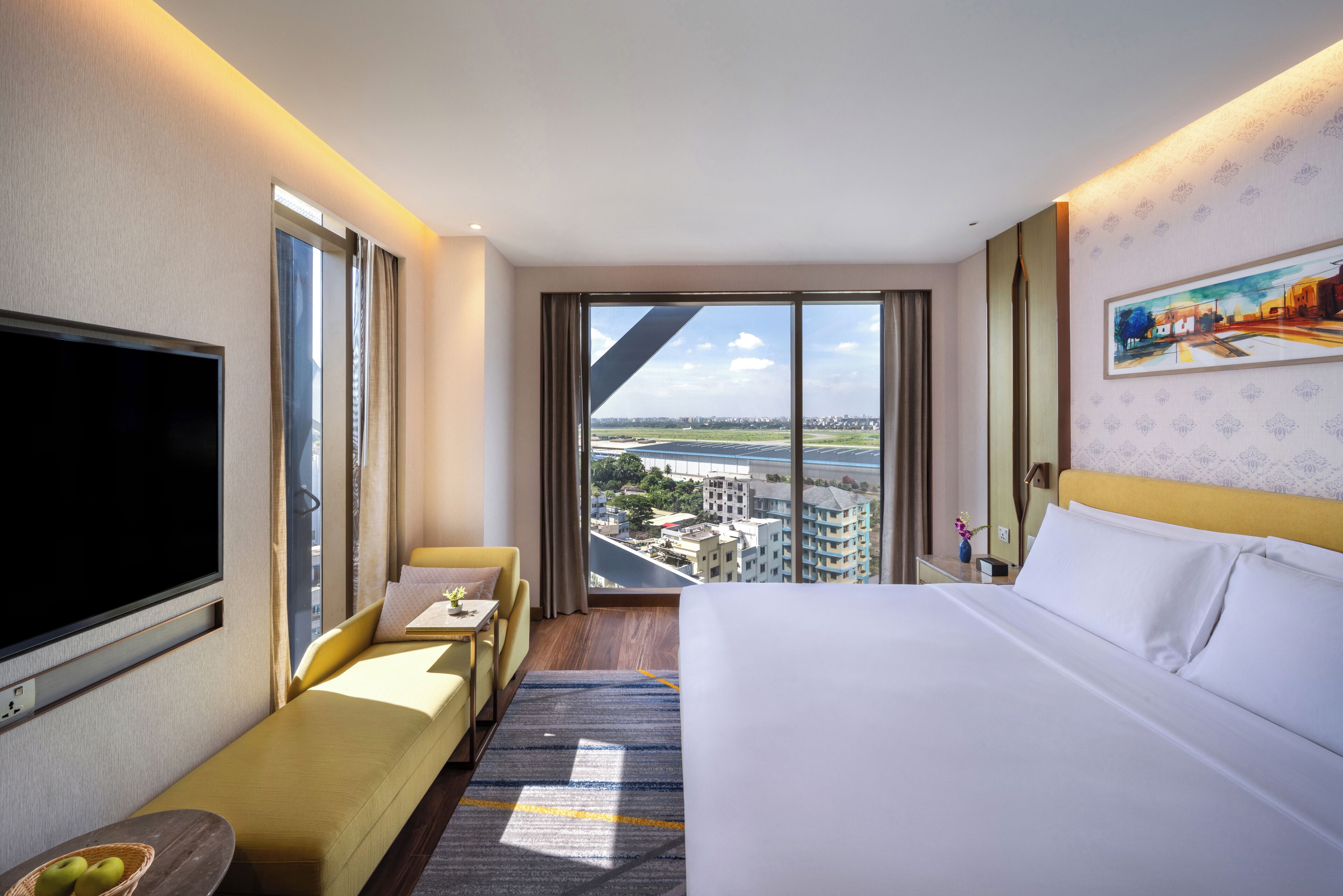 Habitación de hotel con una cama grande, un sofá, un televisor y vistas de la ciudad a través de amplias ventanas.