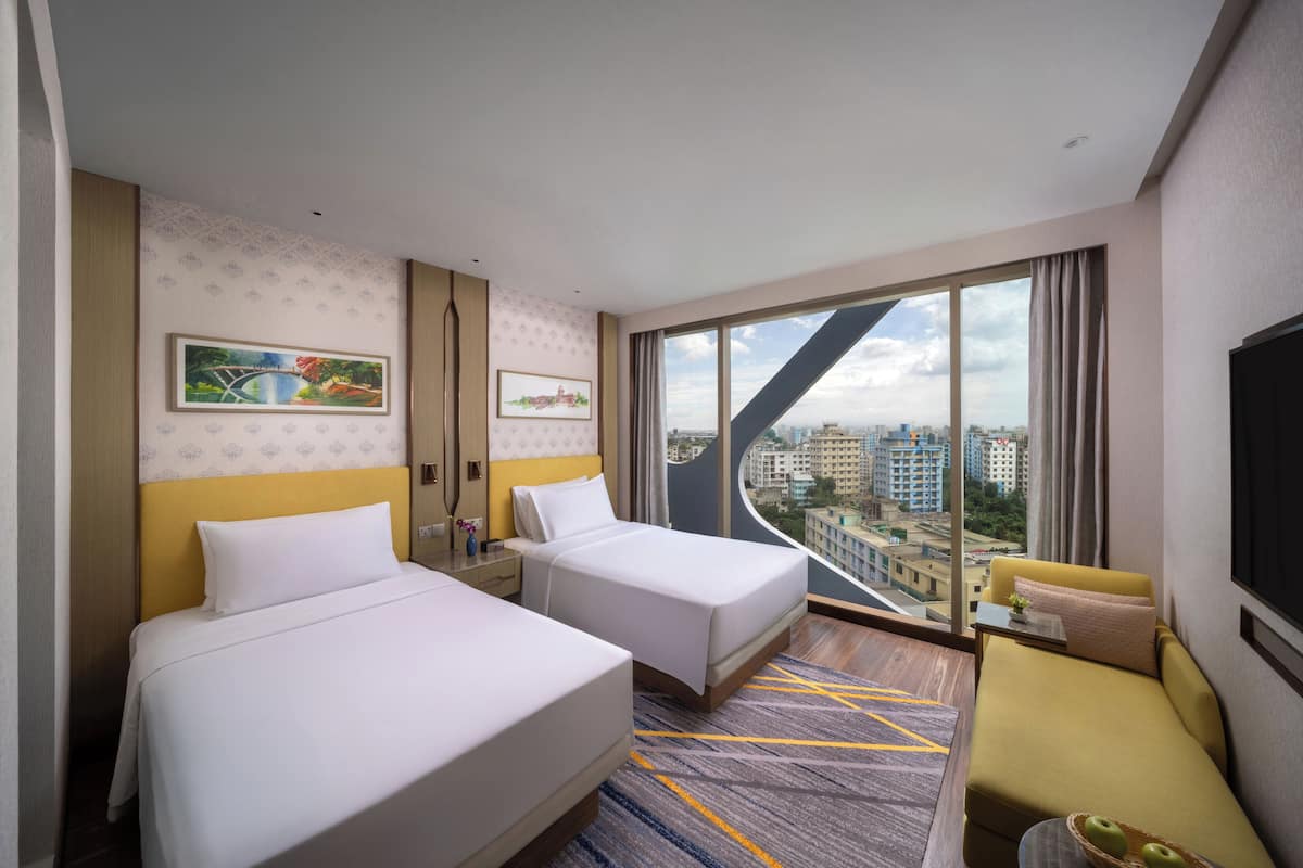 A hotel room with two beds, a sofa, a large window with a city view, and artwork on the walls.
