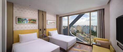 A hotel room with two beds, a sofa, a large window with a city view, and artwork on the walls.
