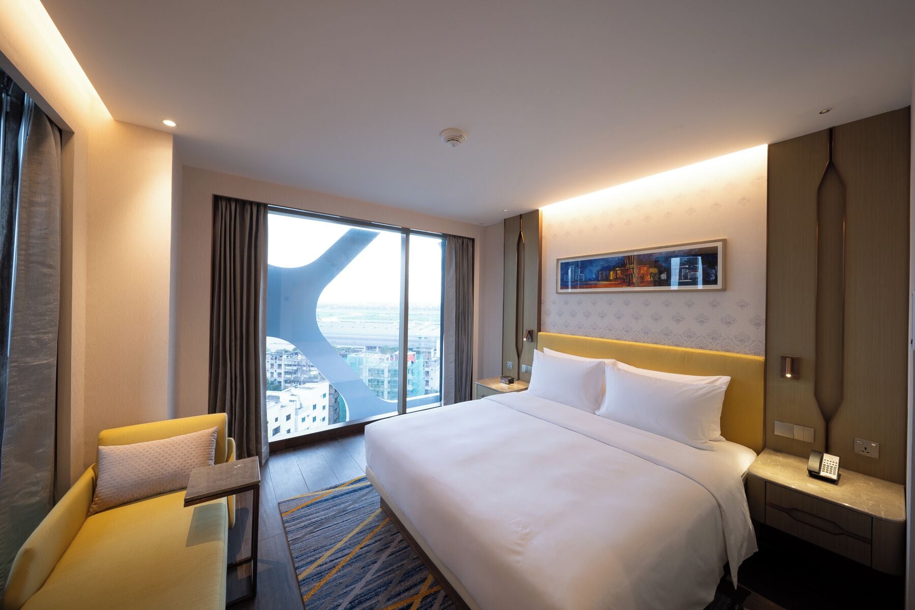 A hotel room with a large bed, a desk, a chair, a nightstand, a painting, and a view of the city.