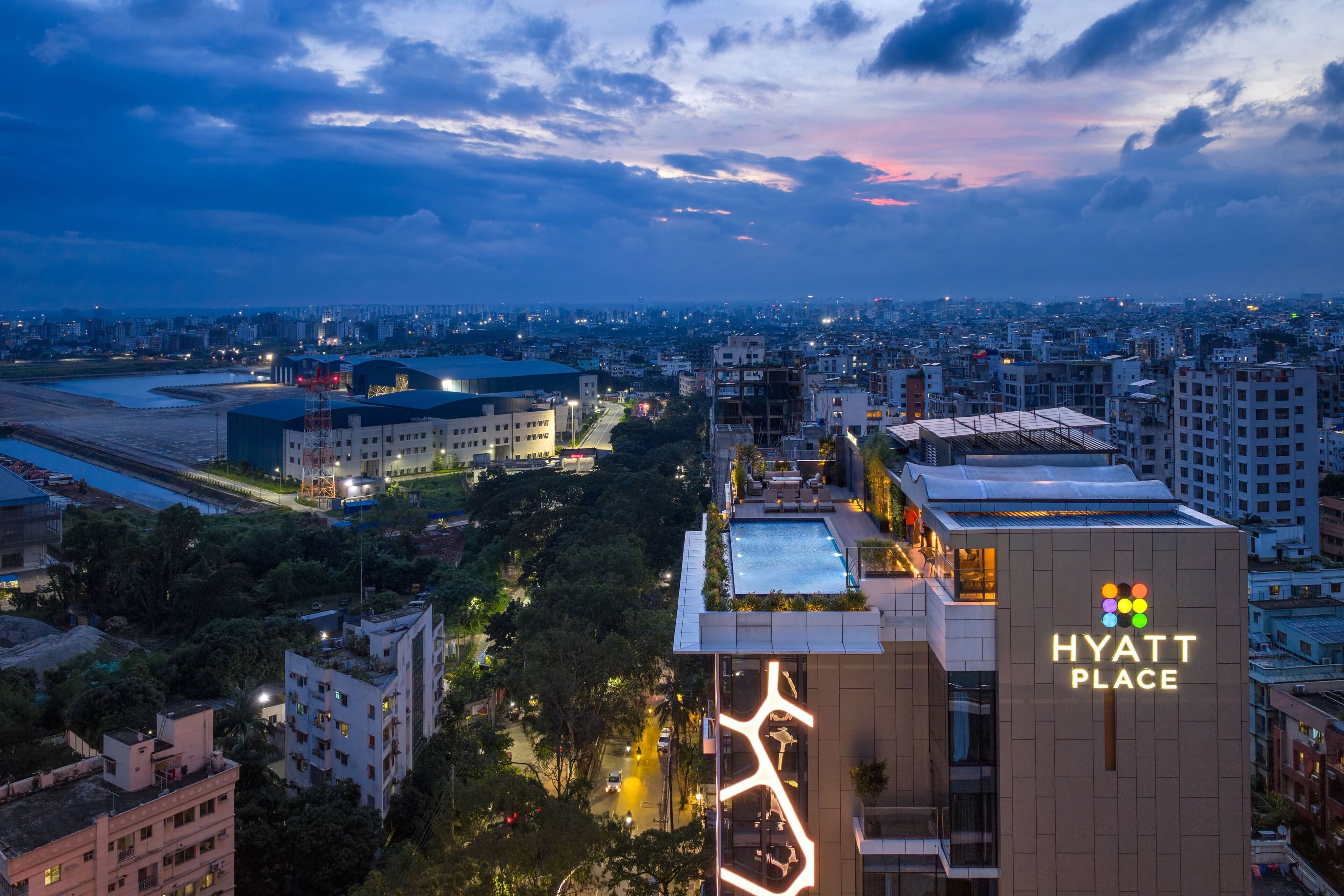 A cityscape at dusk with a prominent Hyatt Place hotel featuring a rooftop pool and a distinctive illuminated sign.