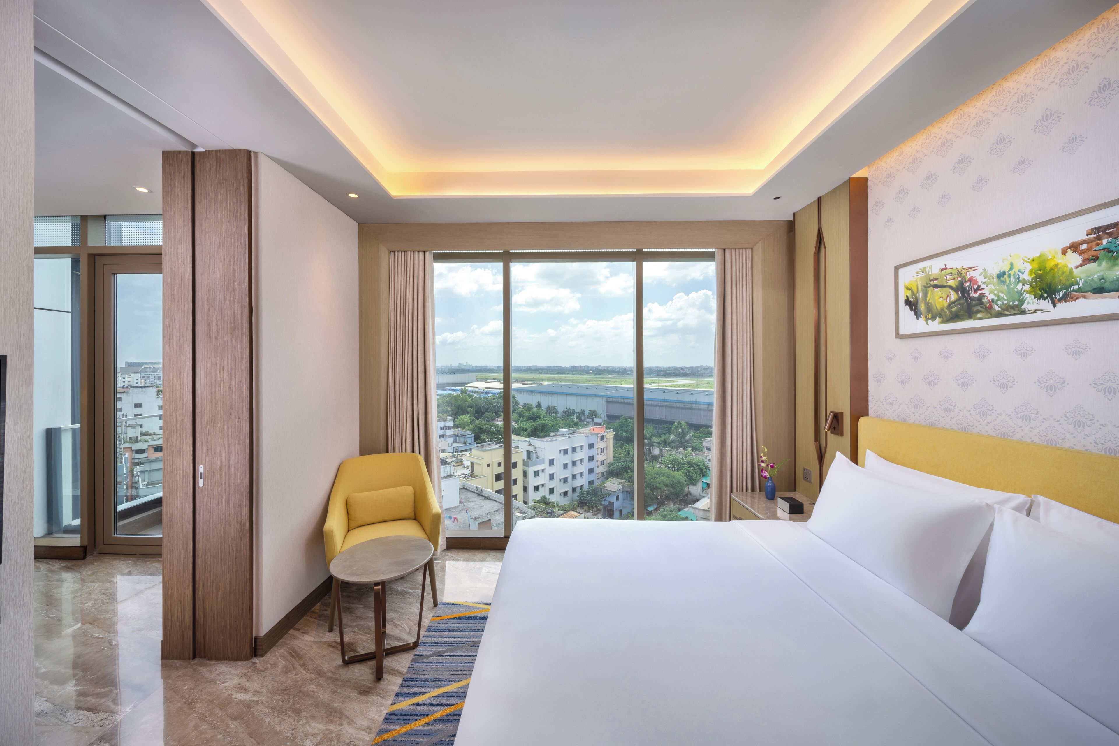 A hotel room with a large bed, a yellow armchair, a view of the city through large windows, and a painting on the wall.