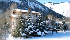 A snow-covered mountain house with a wooden exterior, surrounded by snow-laden trees and a clear sky.