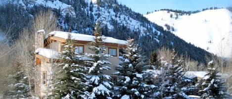 A snow-covered mountain house with a wooden exterior, surrounded by snow-laden trees and a clear sky.