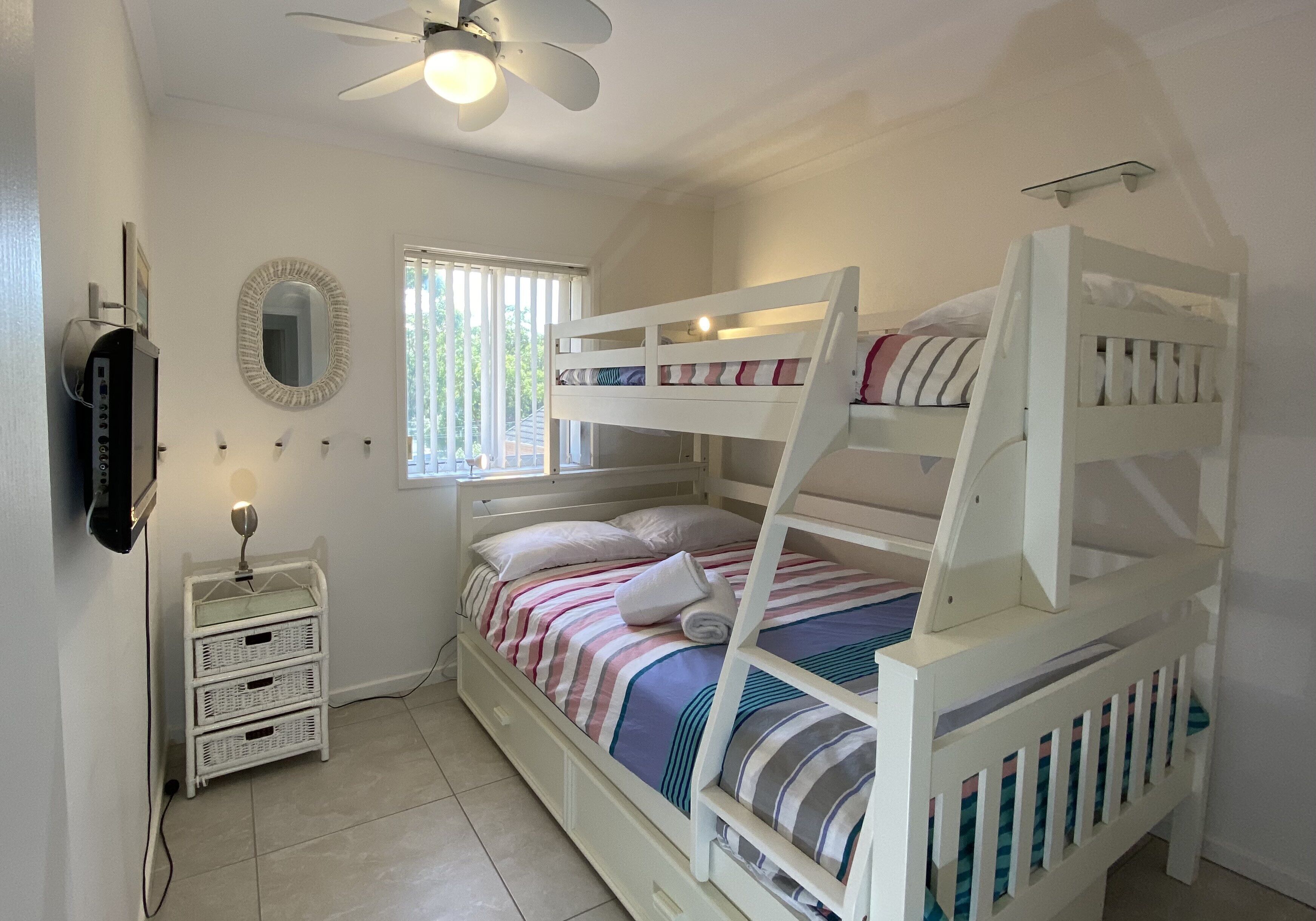 Une chambre avec un lit superposé, un ventilateur de plafond, un miroir et une fenêtre avec des stores.