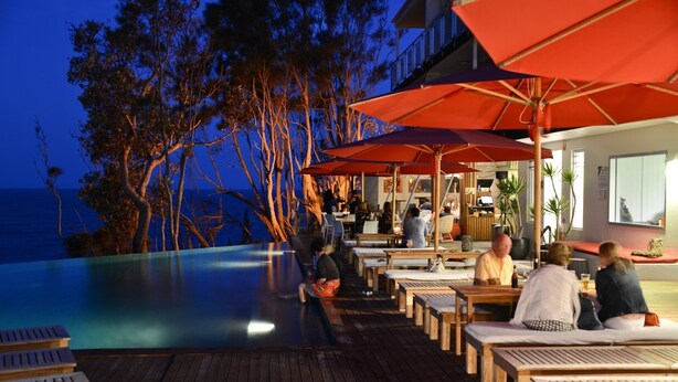 Un espace aménagé au bord de la piscine, avec des chaises longues en bois et des parasols rouges, offrant une vue sur une étendue d’eau et des arbres.