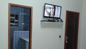 A room with a television on a wall-mounted shelf, a wooden door, and a patterned tile wall. - Acomodação Penedo (Itatiaia)