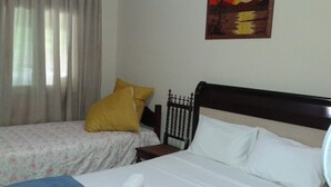 A hotel room with two beds, one with a floral bedspread and the other with a plain white bedspread. There is a painting on the wall depicting a sunset over water. - Acomodação Penedo (Itatiaia)