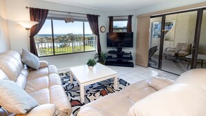 A spacious living room with a large sofa, a coffee table, and a flat-screen TV. There is a balcony with a view of the ocean and palm trees. - CRC 3305 - Third Floor Ocean View Condo (St Augustine)