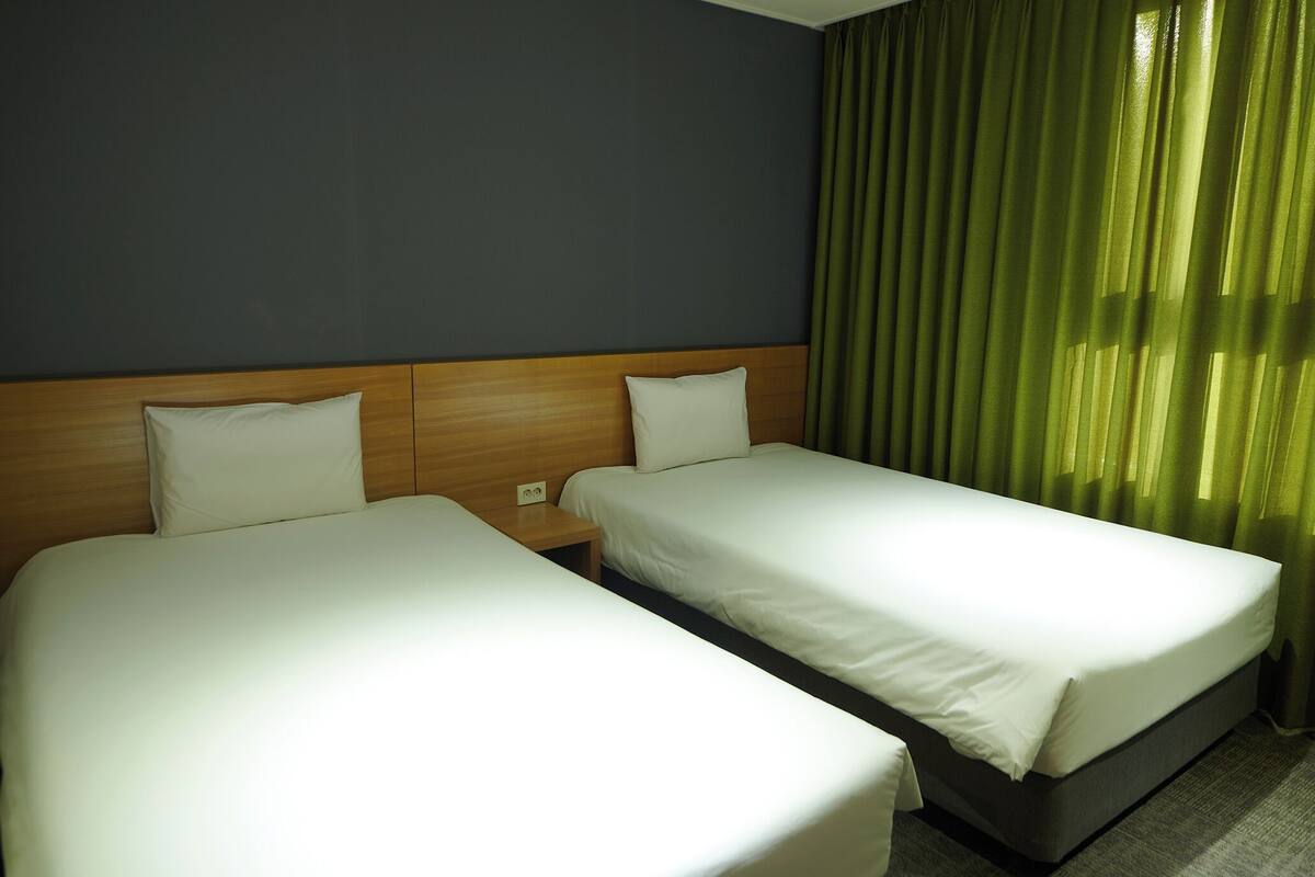 Habitación de hotel con dos camas individuales, cabecero de madera y cortinas verdes.