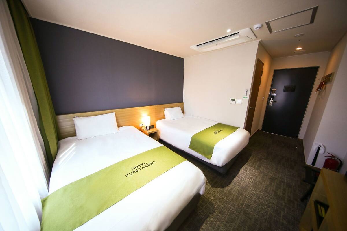 Habitación de hotel con dos camas, una mesita de noche con lámpara, aire acondicionado instalado en la pared, una puerta y un extintor.