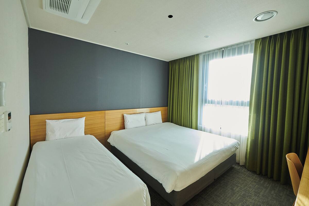 Habitación de hotel con dos camas, cabecero de madera, un ventanal grande con cortinas verdes y suelo alfombrado.