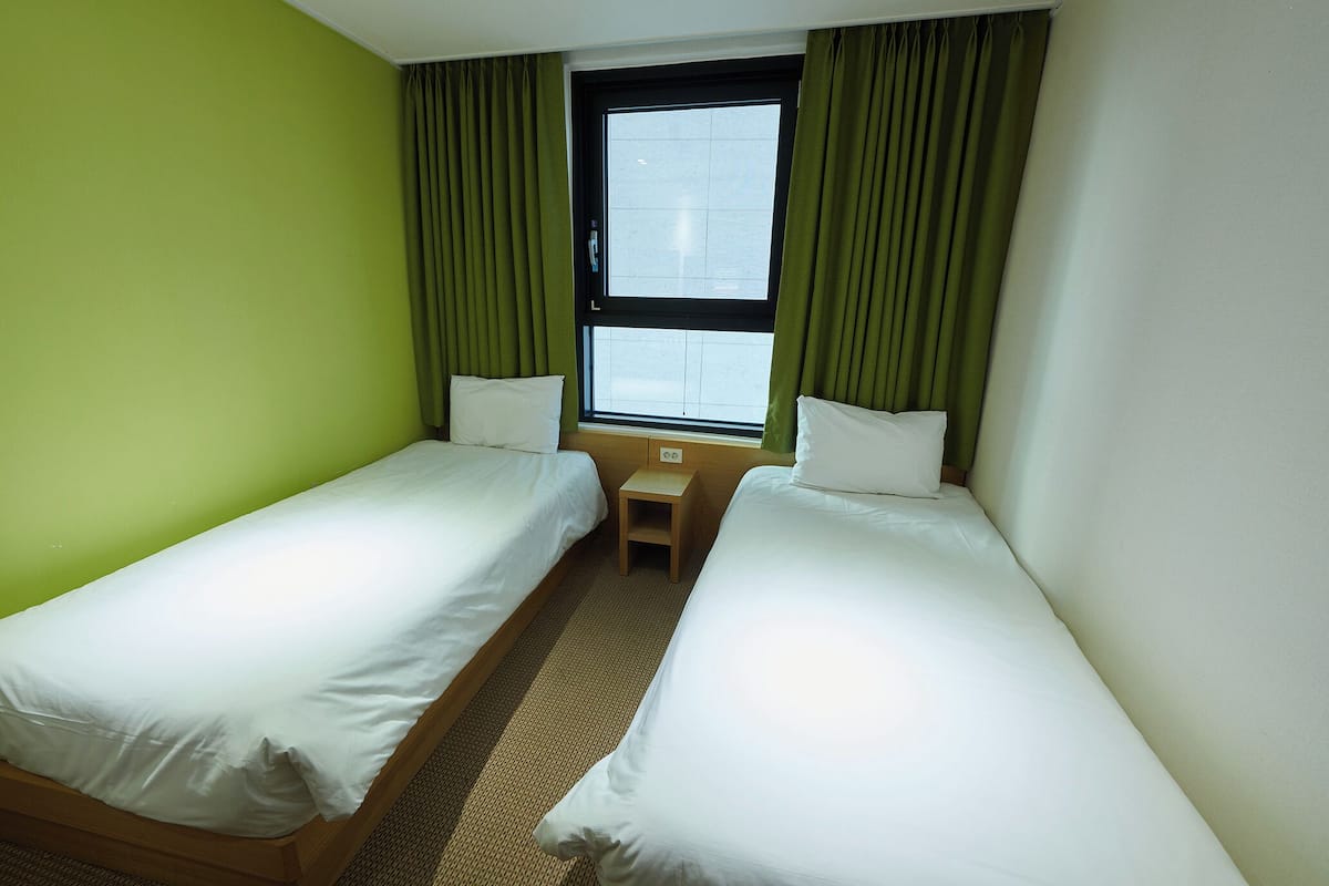 Habitación de hotel con dos camas individuales, una mesita de noche de madera, una ventana con cortinas y una pared verde.