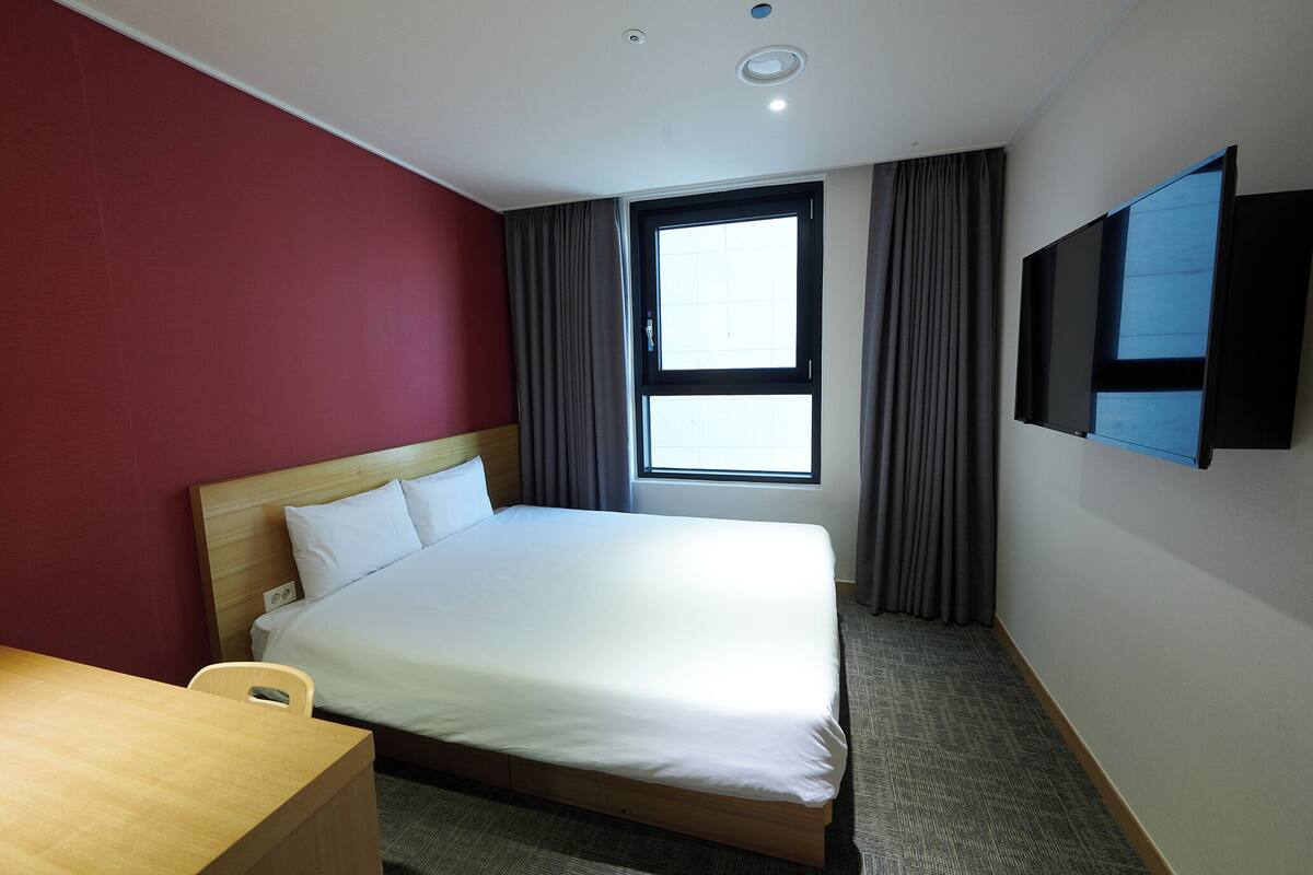 Una habitación de hotel con cama, escritorio, televisión montada en la pared y ventana con cortinas.