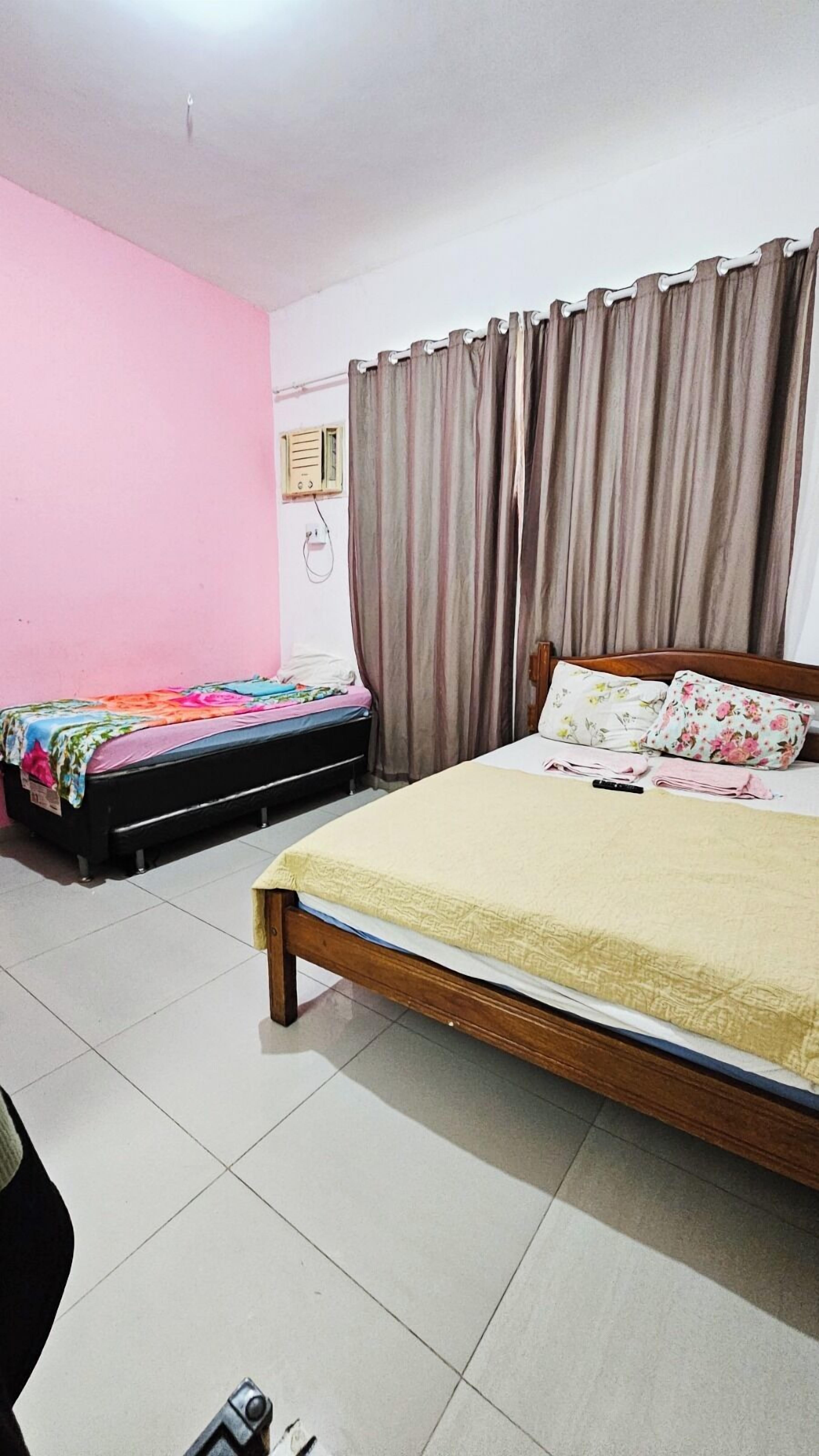 Un dormitorio con dos camas, una más grande y otra más pequeña, pared rosa, ventana con cortinas y un radio montado en la pared.