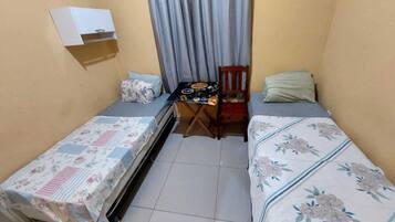 A small, compact room with two single beds, a small table with a chair, and a window with curtains.