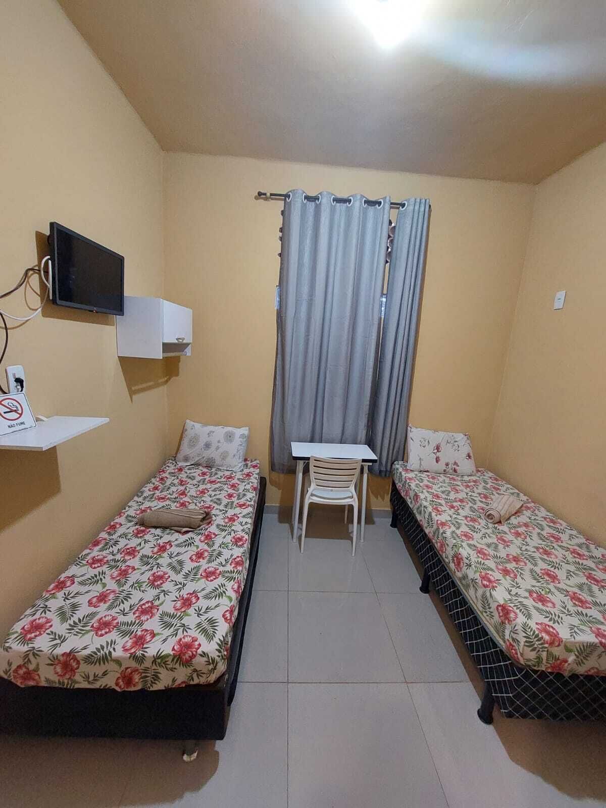 A small, simple room with two single beds, a small table, a chair, a TV, and a curtain.