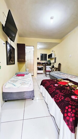 A small, compact room with two single beds, a kitchenette, and a television mounted on the wall. - Pousada Cantinho da Familia (Rio de Janeiro)
