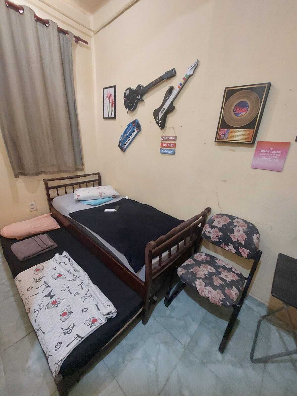 Ein kleines Zimmer mit einem Einzelbett, einem Holzkopfteil, einem stuhl mit Blumenmuster und verschiedenen Wanddekorationen, darunter Gitarren und eine Schallplatte.