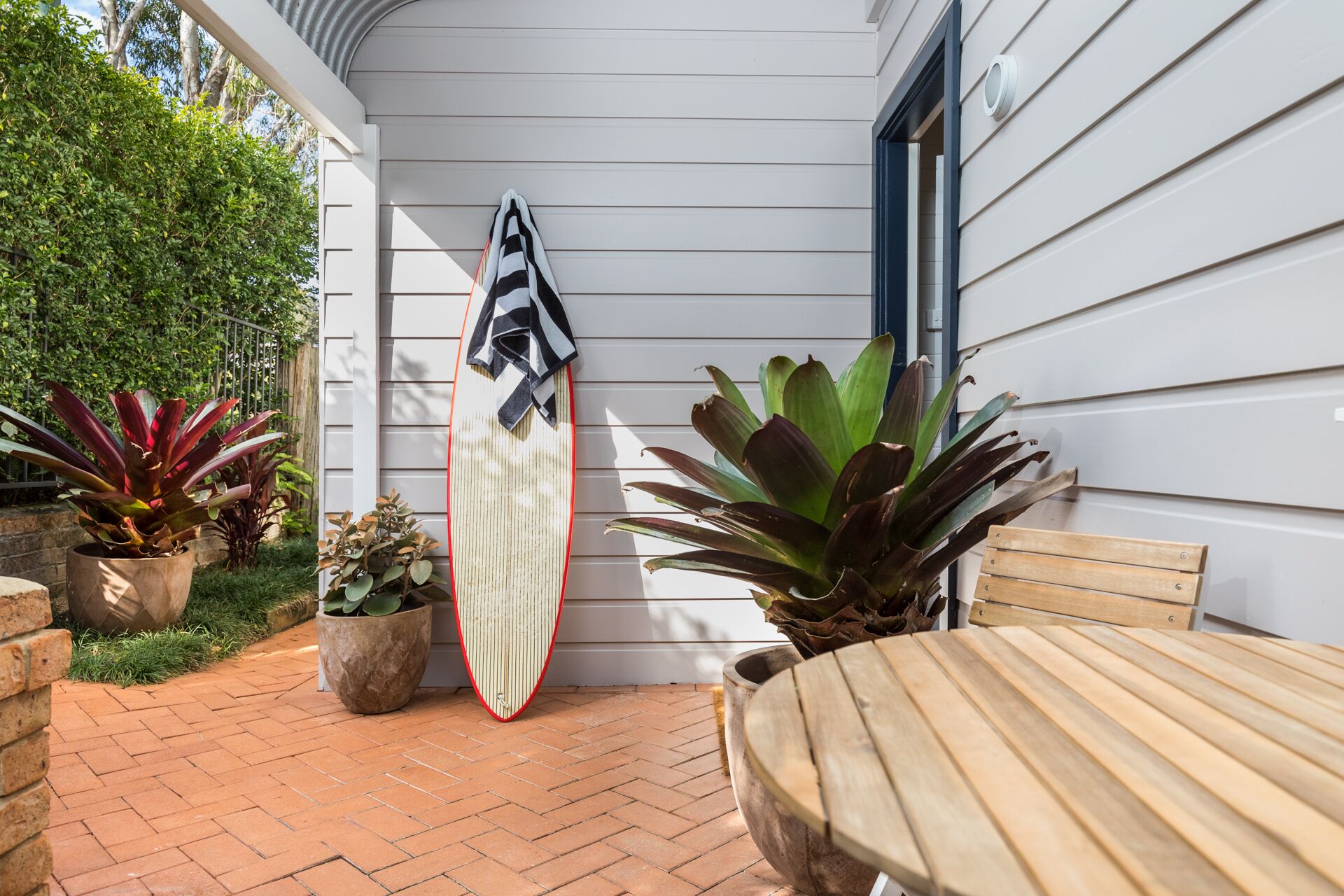A patio area with a surfboard leaning against a house, a wooden table, and various potted plants.