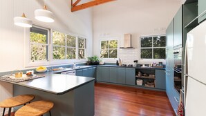 A modern kitchen with a large island, wooden bar stools, and a view of the outdoors through large windows. - Luxurious Jetty House, Coffs Harbour – privacy & tranquility (Coffs Harbour)
