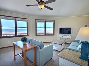 Une pièce dotée d’un ventilateur de plafond, d’une télévision, d’un bureau et d’un canapé. Des fenêtres offrent une vue sur la plage.